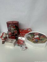 Photo showing large red Coca Cola metal tin with Santa Claus image, rectangular tin, Santa figurine, pack of coasters, cards, stickers, and 3D decorative plate in original box