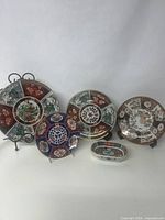 Overview of four Imari decorative plates and one small oval trinket dish grouped together on a neutral background. Plates feature traditional colorful floral and patterned Japanese designs in red, blue, green and gold.