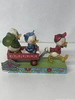 Front and side view of Jim Shore Disney Traditions 'Here Comes Trouble' figurine showing Huey pulling the wagon with Dewey inside and Louie running ahead on a floral patterned base.