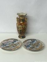 Front view of one 8-inch Satsuma style gold gilded vase with Geisha design and two small matching decorative plates.