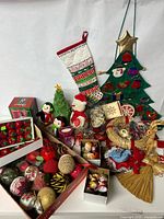 Photo showing a variety of vintage Christmas decorations including a green felt advent calendar shaped like a Christmas tree, Hallmark plush toys (Santa and penguins), a patchwork stocking, many glass ornaments, woven straw ornaments, and small holiday figurines all arranged on a flat surface.
