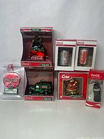 Photo showing full lot including boxed blown glass Coca Cola bottle pack, green glass truck, red logo round ornament, two can-shaped glass ornaments, bear holding can, and small Coke can figurine.