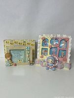 Two vintage Precious Moments baby picture frames displayed side by side with accompanying baby-themed figurines and blocks, showing overall design and condition.