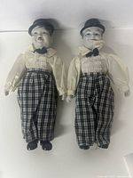 Front view of two porcelain dolls dressed similarly in checked pants, cream shirts, bow ties, and black hats, depicting Stan Laurel and Oliver Hardy.
