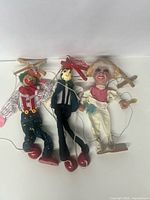 Three handcrafted wooden marionettes including a clown, Pinocchio, and Mexican inspired figure, shown hanging against white background