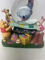 Front view of the musical snow globe showing Pooh, Tigger, Piglet holding patchwork quilt with Eeyore inside globe. Includes picnic basket, honey pots, and flowers on base.