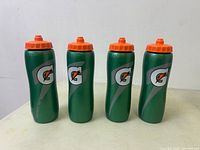 Four green Gatorade bottles with orange caps in a row against white wall, displaying front logos and shape.