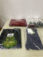 Four pieces of children's clothing, three sweaters including a maroon/red knit, a black Grinch graphic sweater, and a light grey sweater partly visible with a pixelated design. Items are packaged in plastic on a beige surface.