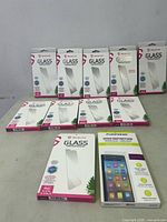 Photo showing multiple packaged tempered glass screen protectors arranged in two rows, mostly from iShieldz brand with white and pink packaging.
