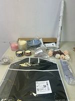 Candles in glass and ceramic holders, small plates, decorative balls, garment bag, and rolled linens on a table.