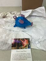 Blue glass flying pig figurine with purple wings, snout, ears and tail shown wrapped in white tissue paper with a description card of Three Crow Glass.