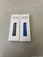 Two unopened i3E EOS LED flashlights in original boxes, one black and one blue, placed side by side on a surface.
