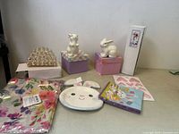 Photo showing two ceramic bunny figurines sitting on pastel decorative boxes, a bunny face plate, Easter-themed napkins, and a crystal candle holder