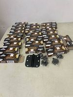 Lot of multiple Zion brand black door hinge packages and loose items on beige surface in rows.