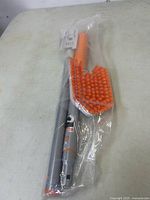 Full view of the new, unopened scrubber set in original plastic packaging, showing extendable handle and orange brush.