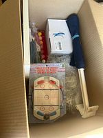 Box containing miniature table hockey game, blue and white umbrella, and white plastic tool with handle packed in a cardboard box with bubble wrap.