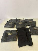Nine black packs labeled 'COMPASS BOOTS' by Saker arranged on a table, showing brand and product clearly with one pack opened revealing a black boot.