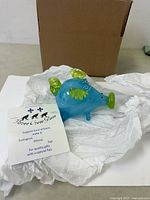 Glass blue pig figurine with green wings and snout wrapped in tissue paper, along with a Three Crow Glass artisan card.