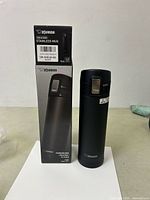 Photo showing black Zojirushi insulated stainless steel mug standing next to its box, displaying the product name and model number SM-KHE48-BA.