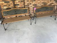 Two wooden outdoor benches with metal lattice backs and wooden slat seats placed side by side on a concrete porch floor against a brick wall.