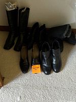 Six pairs of black boots and shoes including tall boots, ankle boots, and leather dress shoes arranged on carpeted floor. Light wear visible.
