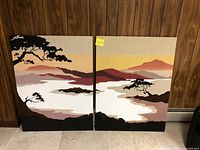 Two burlap landscape wall hanging panels displayed side by side against brown paneling wall on tile floor.