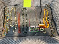 Photo showing 8 assorted colorful and ethnic necklaces showcased on grey knit fabric. Various bead types including glass, wood and possibly bone, different lengths and styles visible.