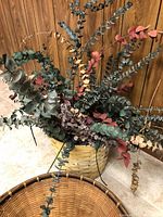 Dried flower arrangement in a woven basket, showing colorful dried eucalyptus stems including green, red, and beige tones.