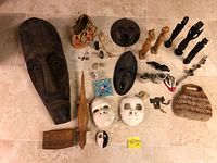 Overview of all items laid out on floor showing masks, wooden statues, ceramic masks, woven purse, carved wooden objects, and Murano glass ashtray.