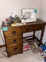 Desk with four drawers on left and one drawer on right, wooden medium-tone finish, vintage metal handles, sewing machine on top, visible wear and dirt.