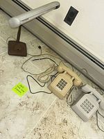 Photo showing vintage desk lamp with a tubular fluorescent bulb and two rotary dial phones, beige and gray, placed on floor next to wall outlet.