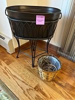 Front view of black metal ice and drink bucket sitting on black metal stand with curved legs and handle loops showing. Smaller round basket placed beside it on wooden floor.