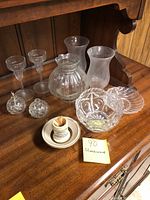 Wide shot of all glassware items on wooden shelf including liqueur glasses, oil lamps, decorative bowls, vases, and toothpick holder.