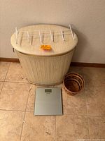 Overview of oval wicker hamper with white hook rack on top, small round wicker trash can, and Taylor glass top digital scale on tile floor.