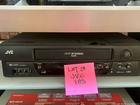 Front view of the black JVC VHS Hi-Fi stereo VCR with tape slot and control buttons visible, with a pink sticky note labeled 'LOT 18 JVC VHS'.