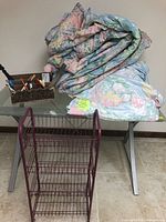 King size floral comforter folded on table with matching floral sheets or pillowcases, small office organizer with pens and markers, and burgundy metal rolling cart on floor.