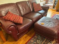 Full view of dark cranberry leather sofa with two striped pillows and matching ottoman on a hardwood floor with area rugs