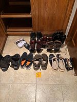 Wide view of men's shoes lot including dress shoes, slip-on shoes, boots, sandals, and slippers arranged on floor and shoe rack.