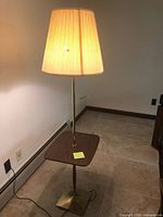 Full view of the lamp standing against a wall, showing cream-colored shade, brass pole and base, and attached wood-tone table surface.