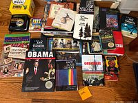 Photo showing a collection of books including 'Obama: The Historic Presidency,' 'Like a Lily Among Thorns,' 'To Kill a Mockingbird,' Dolly Parton biography, and others on a wooden floor.