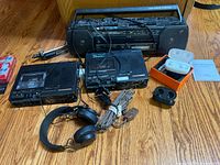 Overview of music electronics lot including cassette players, headphones, earbuds, antenna, and tapes on wood floor