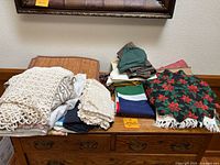 View of assorted folded placemats, tablecloths, napkins, and runners stacked on wooden dresser. Includes crocheted and lace linens and woven textiles.