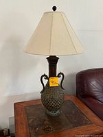 Full view of the lamp sitting on a wooden side table with a beige lampshade and amphora-style base with curved handles.