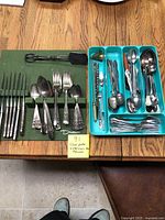 Two trays of flatware on a table showing a Drew silverplate flatware set on a green mat and a turquoise tray with varying stainless steel flatware items including spoons, forks, tongs, chopsticks, and a candle snuffer.