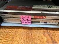 Sony DVD recorder RDR-GX315 and separate Sony DVD player stacked on a wooden floor, with a pink sticky note identifying them as lot 19.