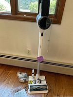 Full view of white Tineco cordless vacuum in upright position with main floor brush head on wooden floor near window.