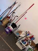 Photo showing walking sticks, a mop, brooms, dusting brushes, white storage racks with various items, black bucket, and some wooden boxes.