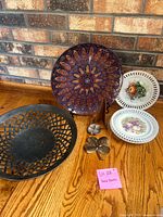 Photo showing a large colorful decorative plate, two smaller white ceramic plates with fruit and floral patterns, a large black metal lattice-style plate, and three shamrock-shaped metal trinkets on a wooden floor.