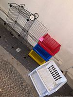 Metal wire rolling cart with three tiers and wheels, red, blue, and yellow plastic stackable organizers next to it on floor