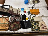 Full view of nine different women's purses of various styles and colors, displayed on a bed with seven beauty items visible behind them. Bags include purses made of leather, fabric, tapestry, and woven materials.